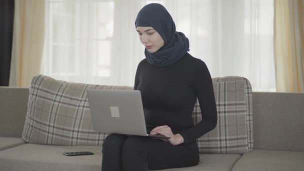 Young smiling successful confident girl in balck clothes and beautiful headdress working with her laptop sitting on the sofa at home. Young asian Muslim woman in headscarf surfing the web - Video