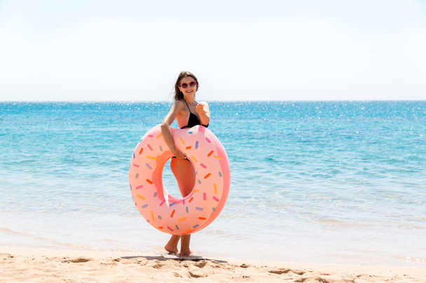 woman calls to swim in the sea and waves her hand. Girl relaxing with donut on the beach and playing with inflatable ring. Summer holidays and vacation concept - Fotografie, Obrázek