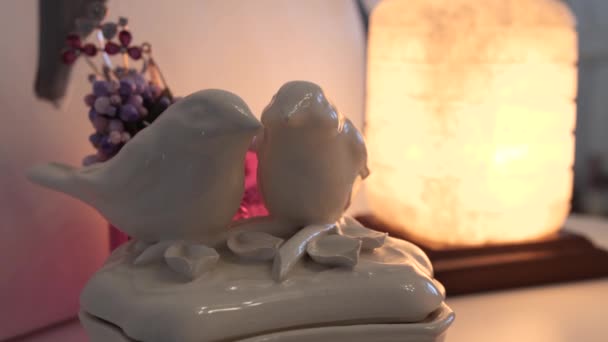 Πορσελάνη αγαλματάκι με τη μορφή δύο αγαπησιών πουλιών που στέκονται στο ράφι με φόντο τη λάμπα στο εσωτερικό του σπιτιού. 4K αργή mo - Πλάνα, βίντεο