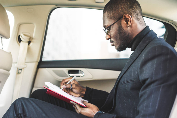 A young business man in a suit is sitting in the back seat of a expensive car with a notebook. Business negotiations. - Foto, immagini