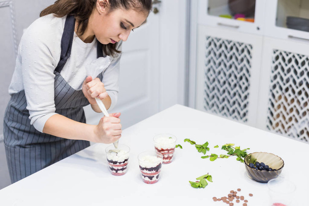 A confectioner prepares a trifle in three cups. Desserts are on the white table in the kitchen. The concept of homemade pastry, cooking cakes. - Photo, image