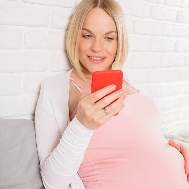 Happy smiling pregnant woman lies in bed and looks at mobile phone - Foto, Bild
