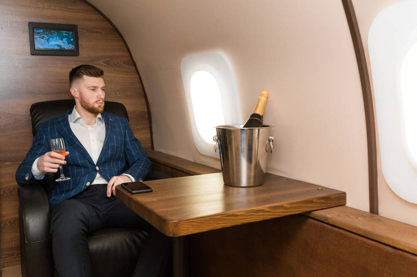 A young successful businessman in an expensive suit sits in the chair of a private jet with a glass of champagne in his hand and looks out the window - Zdjęcie, obraz