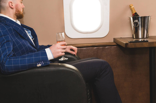A young successful businessman in an expensive suit sits in the chair of a private jet with a glass of champagne in his hand and looks out the window - Φωτογραφία, εικόνα
