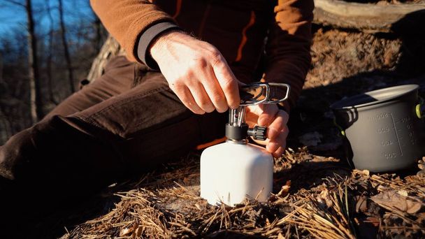 The theme of tourism is hiking and traveling in nature. Hands A Caucasian man uses equipping to cook food outside. A tourist installs a stove burner on a balloon cartridge with gas for boiling water - 写真・画像