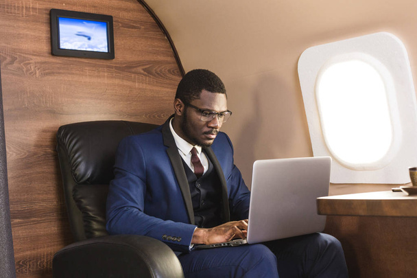 Attractive and successful African American businessman with glasses working on a laptop while sitting in the chair of his private jet. - Foto, imagen