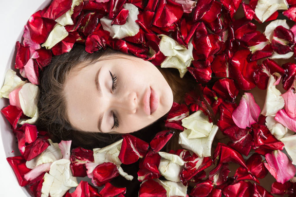 A young attractive girl takes a bathroom with flower petals and relaxes against the background of a beautiful light interior. Spa treatments for beauty and health with skin care - Photo, Image