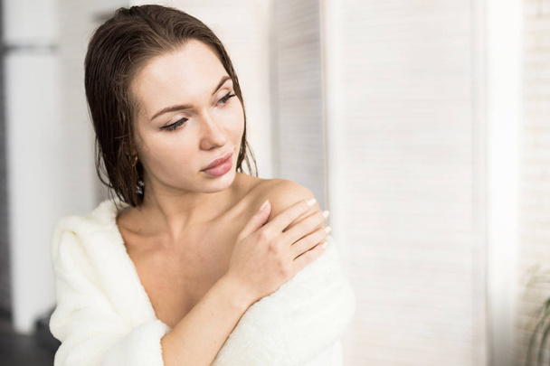 Attractive young girl in a white bathrobe applies cream to the skin while sitting in the interior of the bathroom and doing morning procedures. Skin and body care - Photo, image