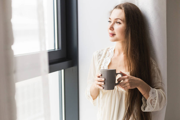 Attraktives junges Mädchen in Nachthemd, das mit einem Becher Kaffee am Fenster steht - Foto, Bild