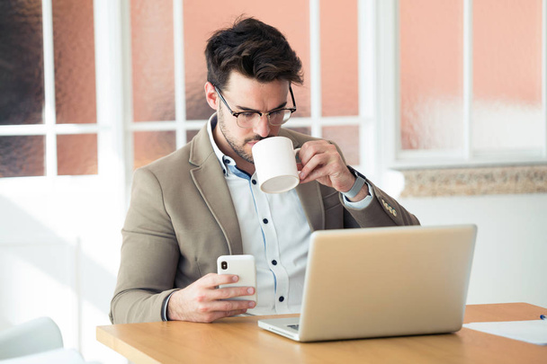 Handsome young business man using his smartphone while drinking coffee in front of laptop in the office. - Foto, imagen