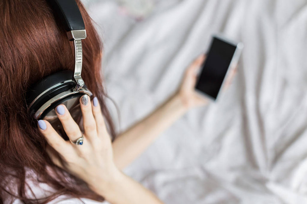Attractive young brunette woman sits in the morning in her bed with headphones and a smartphone and listens to music in a good mood - Valokuva, kuva