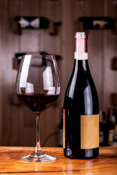 Γυαλί και μπουκάλι με νόστιμο κόκκινο κρασί σε ξύλινο τραπέζι. Στο παρασκήνιο κόκκινα μπουκάλια κρασιού στοιβάζονται σε ξύλινα ράφια shot με περιορισμένο βάθος πεδίου. - Φωτογραφία, εικόνα