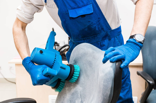 Young man in workwear and rubber gloves cleans the office chair with professional equipment. - Fotoğraf, Görsel