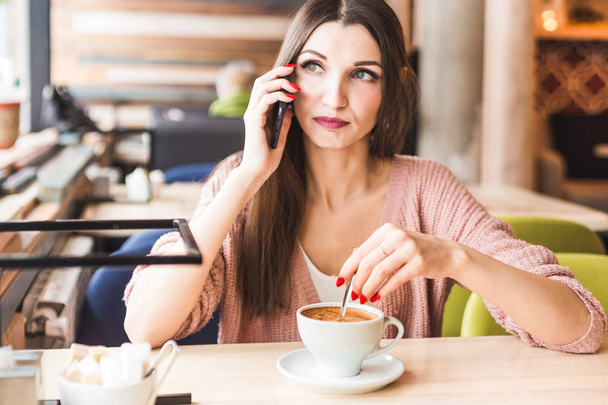 Kaunis nuori nainen istuu pöydässä kahvilassa kupin kahvia ja puhuu puhelimessa.
 - Valokuva, kuva