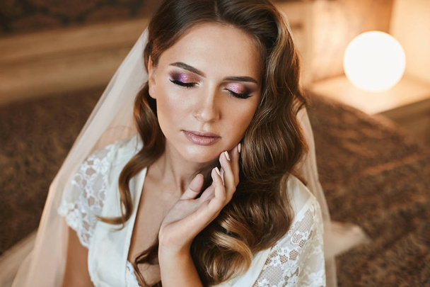 Beautiful brown-haired woman with wedding hairstyle and bright makeup sits with closed eyes on the bed - Photo, Image