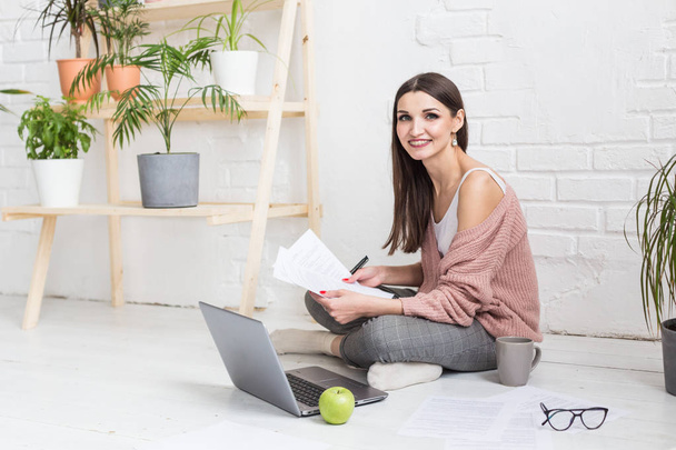 A young happy woman sits on the floor in a bright apartment or office interior and works at a laptop, freelancer girl at work, the concept of distance learning students, paperwork and online learning - Fotografie, Obrázek