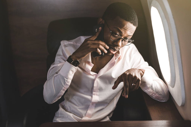 Young attractive and successful African American businessman with glassies talking on the phone and working while sitting in the chair of his private business plane - Foto, immagini