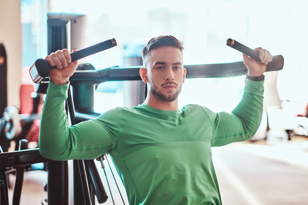 Nachdenklicher junger Bodybuilder im grünen Hemd macht Übungen an Trainingsgeräten - Foto, Bild