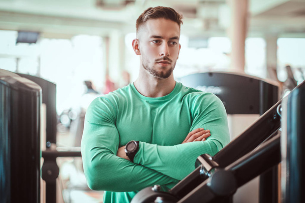 Jovem fisiculturista pensativo em camisa verde está de pé perto do aparelho de treinamento com as mãos cruzadas
 - Foto, Imagem