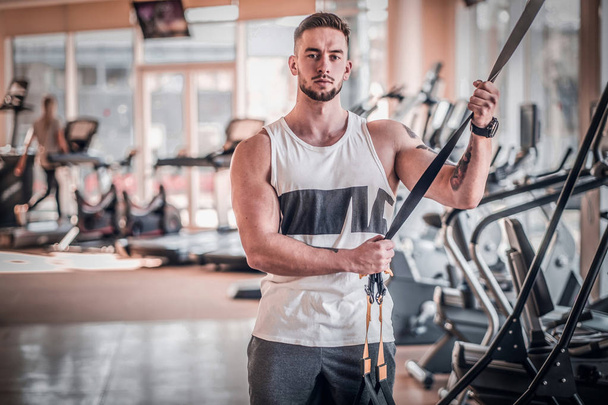 Atraente jovem fisiculturista está posando ao lado do aparelho de treinamento no ginásio ensolarado
 - Foto, Imagem