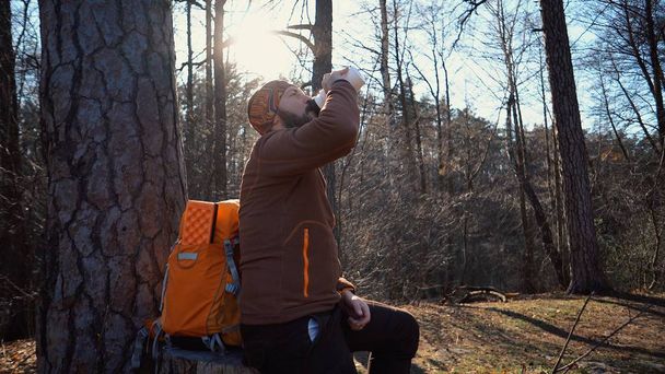 A young tired Caucasian man carries a rest break with a backpack sitting and drinking water on a tree in a nature forest during hikes, trips, vacations, on vacation, outdoor lifestyle freedom concept - Photo, Image