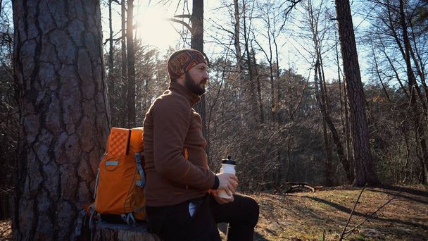 Un joven hombre caucásico cansado lleva un descanso con una mochila sentada y bebiendo agua en un árbol en un bosque natural durante caminatas, viajes, vacaciones, vacaciones, concepto de libertad de estilo de vida al aire libre
 - Foto, Imagen