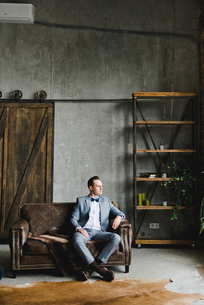 Porträt eines Mannes. Der Bräutigam im grauen Anzug, weißem Hemd und Fliege sitzt auf einem braunen Ledersofa. Zimmer im Loft-Stil. - Foto, Bild