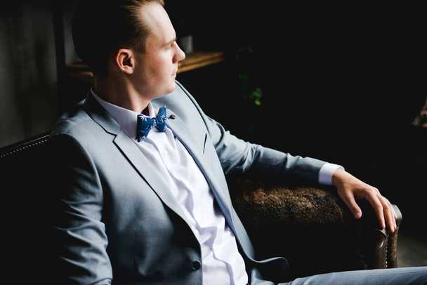 Egy ember portréja. A vőlegény, a szürke öltönyt, fehér inget és egy csokornyakkendő ül egy barna bőr kanapé. Loft stílusú szoba. - Fotó, kép