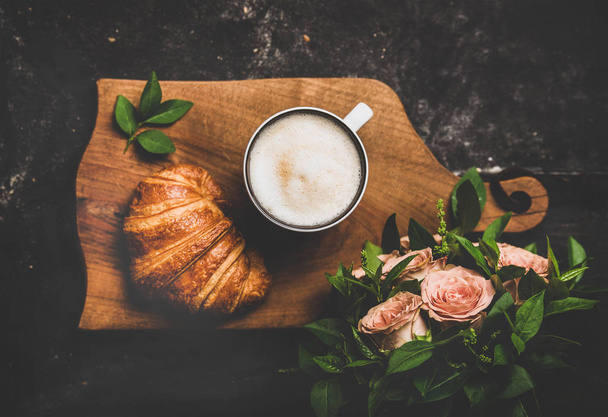 Coffee break. Flat-lay of cup of hot cappuccino with croissant and bouquet of pink flowers over black shabby background, top view. Spring mood - Foto, immagini