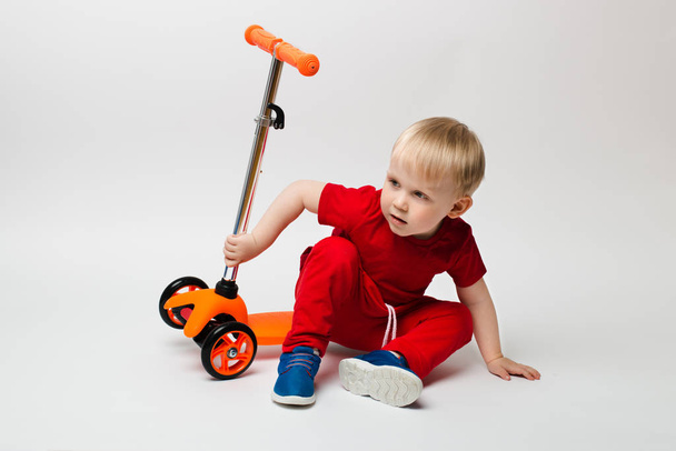 Cute Toddler child in a red t-shirt fell from a scooter and sits, shooting in the studio on a white background - Photo, Image