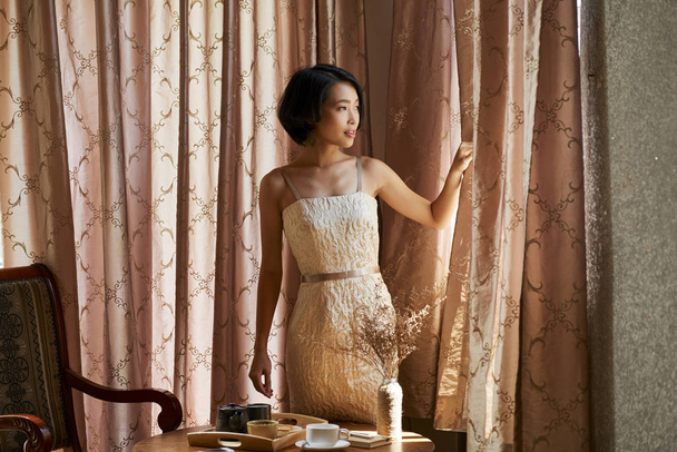 素敵な若いアジアの女性は、リビングルームでカーテンを開け、窓を通して見て - 写真・画像