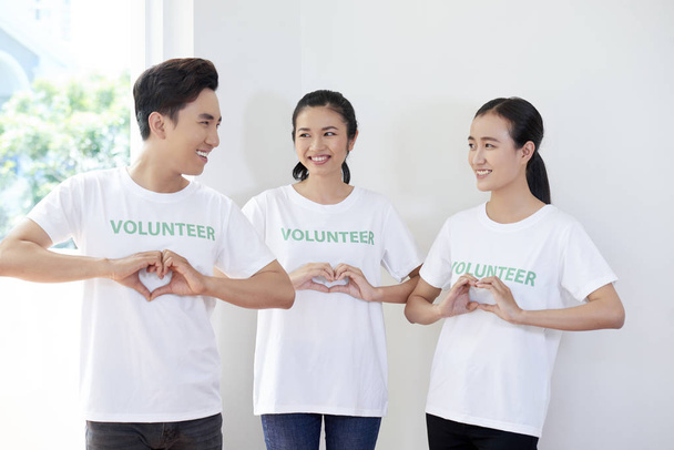 Team van jonge vrijwilligers die in witte t-shirts staan en een hart vorm geven vanuit hun handen en glimlachen - Foto, afbeelding