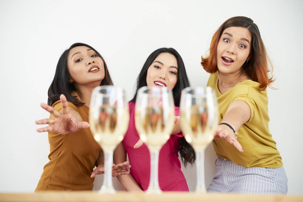 Tres mujeres jóvenes de pie y buscando copas de champán que están sobre la mesa
 - Foto, Imagen