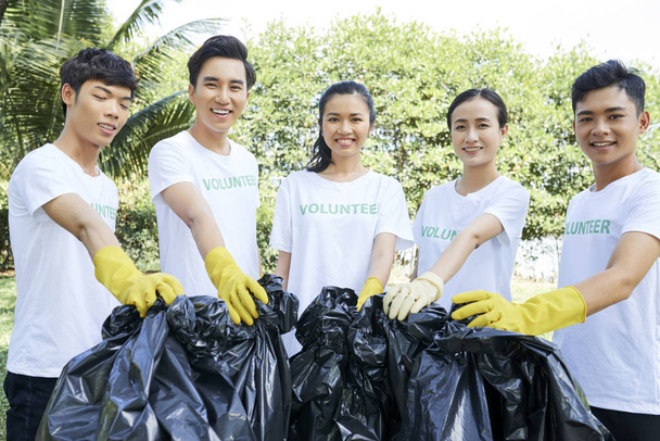 Portrét asijské dobrovolnické skupiny, stojící pohromadě v ochranných rukavicích a držící pytle s odpadky, s úsměvem venku na kameře - Fotografie, Obrázek