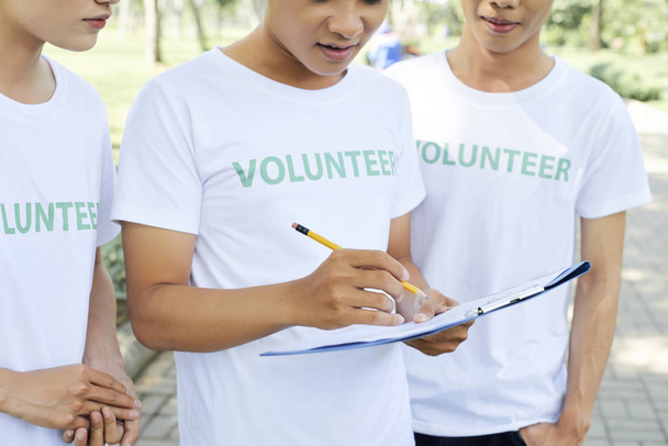 Primer plano del grupo de voluntarios con camisetas blancas haciendo notas con lápiz en el documento mientras trabajan juntos al aire libre
 - Foto, Imagen