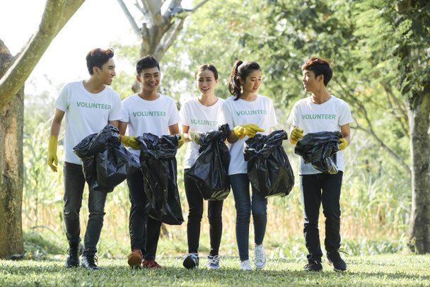 Группа молодых азиатских добровольцев, прогуливающихся в парке с мешками для мусора, они убирают территорию вокруг
 - Фото, изображение