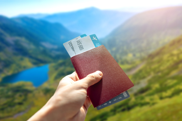 Hand holding a passport and two air tickets on a blue background of mountains. Travel and vacation in beautiful places - Photo, Image