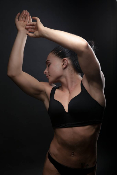 photo of a young athletic woman sports build in a swimsuit. Theme sports and bodybuilding. A woman on a black background hands up - Фото, зображення