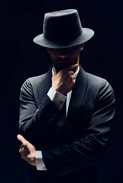 Handsome thoughtful man in black suit and hat touching chin isolated on dark background                        - Valokuva, kuva