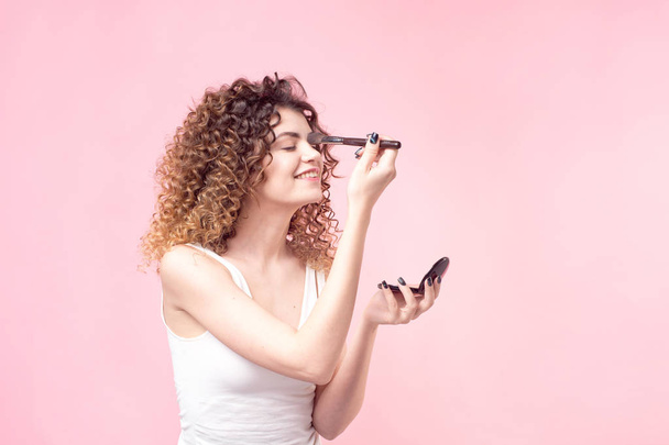 Profile of cheerful young female is standing with hand mirror and powdering her nose using professional brush isolated on pink background - Photo, image