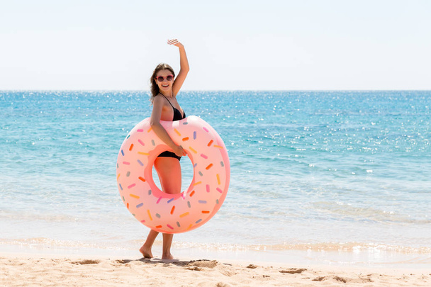 mulher chama para nadar no mar e acena a mão. Menina relaxante com donut na praia e brincando com anel inflável. Conceito de férias e férias de verão
 - Foto, Imagem