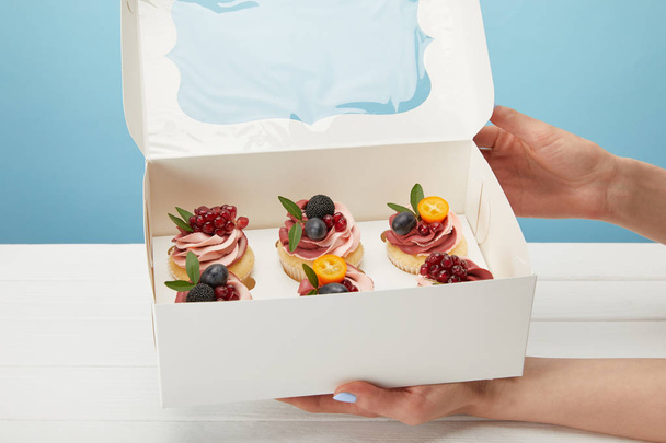 partial view of woman holding box with cupcakes isolated on blue - Fotó, kép