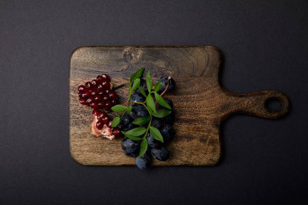 top view of grapes and garnet on wooden cutting board on black surface - Photo, Image