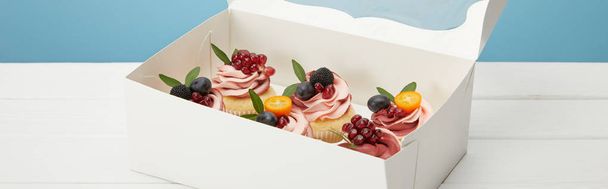 Panoramaaufnahme von Cupcakes mit Beeren und Früchten in Schachtel auf weißer Oberfläche isoliert auf blauer - Foto, Bild
