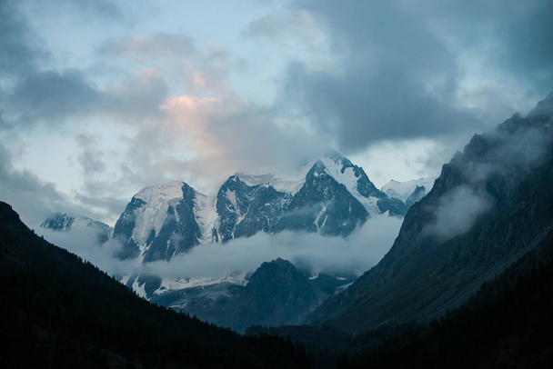 Low cloud before huge glacier. Giant snowy rocky mountains under cloudy sky. Thick mist in mountains above forest at early morning. Impenetrable fog. Dark atmospheric landscape. Tranquil atmosphere. - Photo, Image