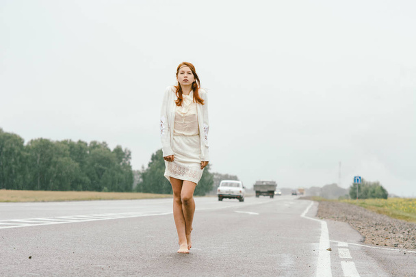 Рудоволоса дівчина в білому одязі ходить босоніж вздовж доріжки в дощову погоду
. - Фото, зображення