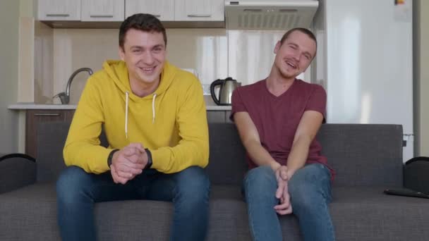 Two men are sitting on the couch and smiling. Young guys are talking, turn away their faces and laugh. A kitchen on the background. 4K footage. - Footage, Video