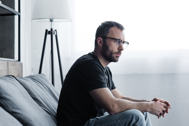 thoughtful handsome man in glasses sitting on grey sofa and looking away - Foto, imagen