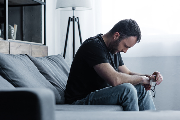 depressed man holding glasses while sitting on grey sofa at home - Photo, Image