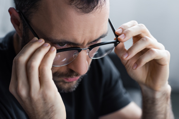 handsome serious unshaven man looking away and touching glasses  - Фото, изображение